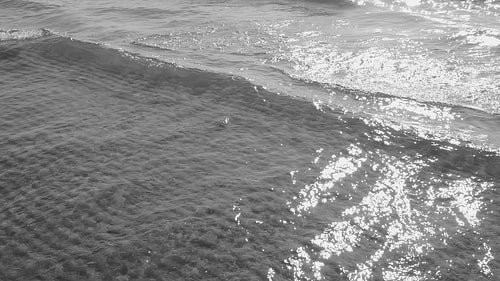 Tracking slow motion sunlit lake wave in shallow water. Rippled sand. Black and white. HD stock video.